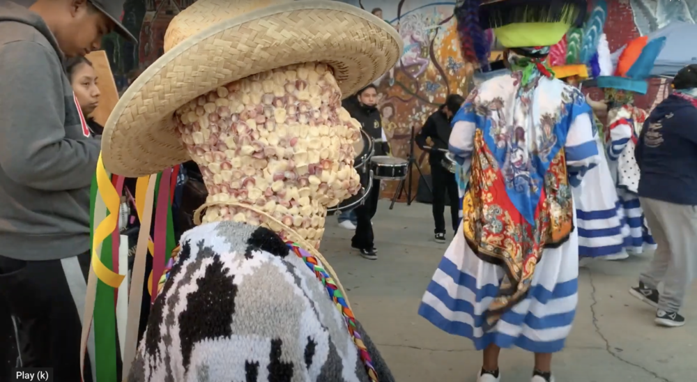 a figure made of corn kernels wearing a cowboy hat and poncho is in from of colorfully dressed traditional dancers