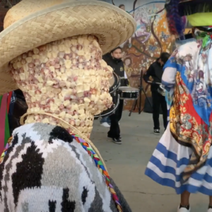 a figure made of corn kernels wearing a cowboy hat and poncho is in from of colorfully dressed traditional dancers