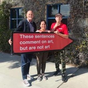 Artist Yumi Roth stands with GCAC Director John Spiak and Aaarrow sign spinner holding red arrow sign with white text by artist Sol Lewitt that reads "These sentences comment on art, but are not art"