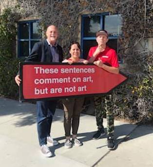 Artist Yumi Roth stands with GCAC Director John Spiak and Aaarrow sign spinner holding red arrow sign with white text by artist Sol Lewitt that reads "These sentences comment on art, but are not art"