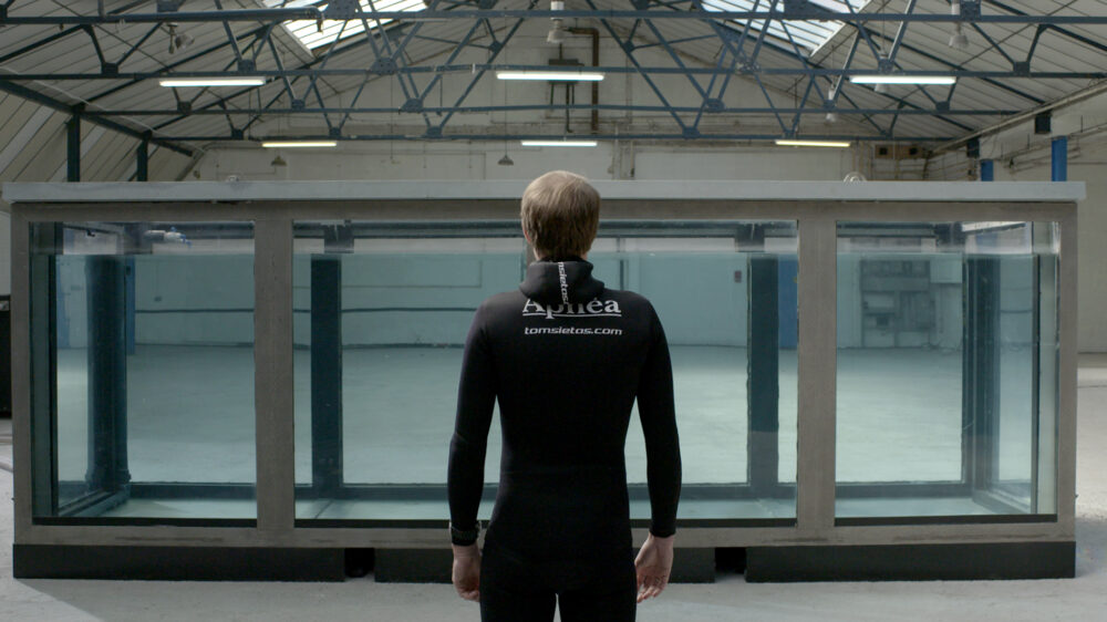 Man in wetsuit standing in front of tank filled with water.