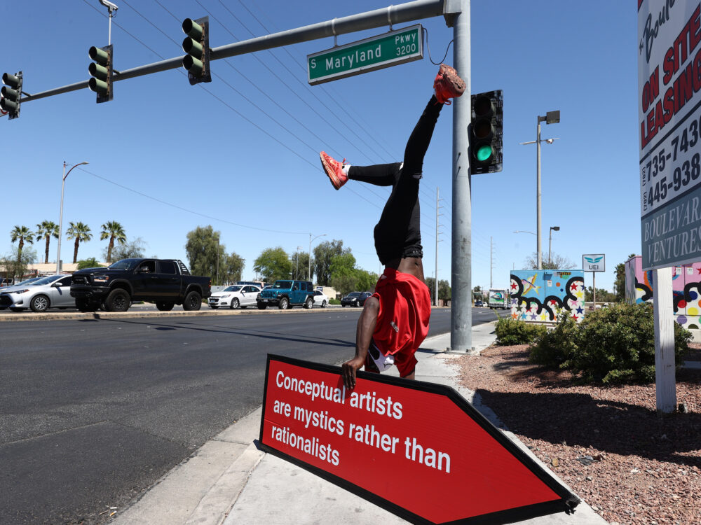 a street corner with an individual sign spinner doing a handstand on a red and white arrow sign