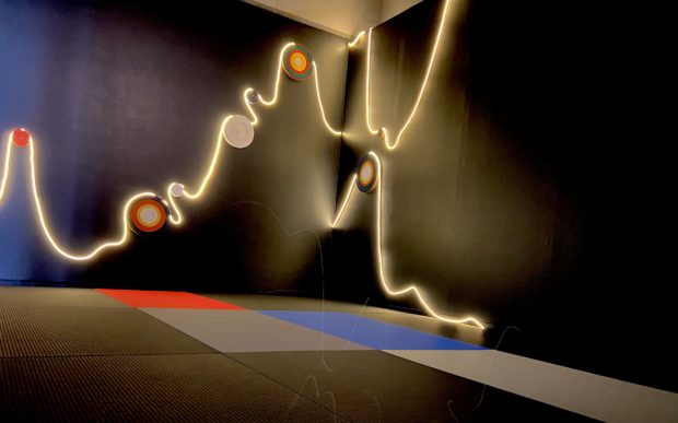 LED light string hanging in fluid like motion on a black wall, with black, red, blue and gray matts on floor