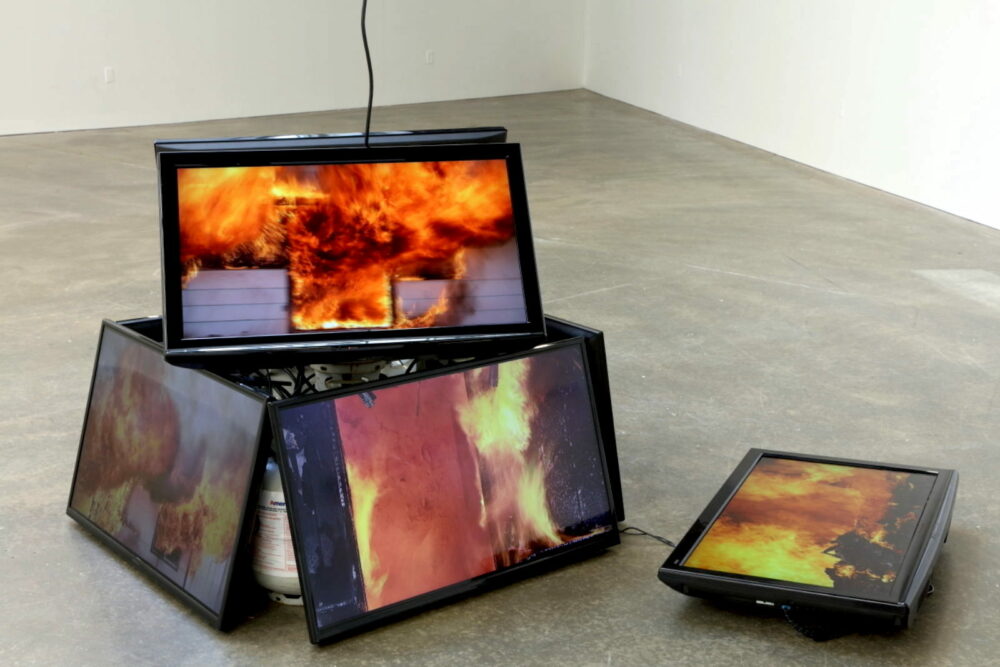 Multiple TV monitors installation on the floor, projecting images of houses on fire.