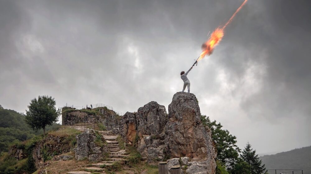 women in white outfit standing on rock formation in the right center of image shooting a flaming arrow into the sky.