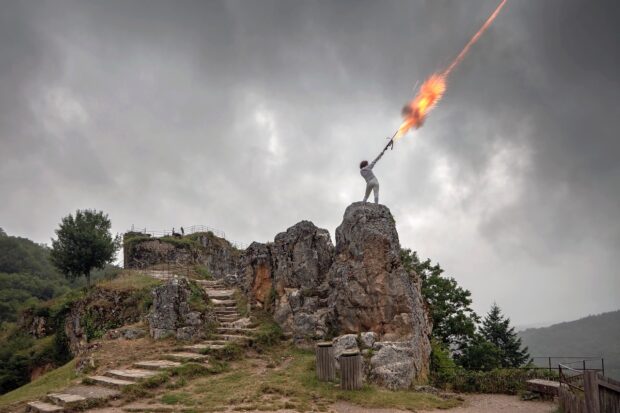 Woman in white outfit to right center of image stings on a natural rock formation and shots a fire arrow into the sky.