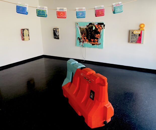 colorful ice coolers hang across a rope toward the top of the image, with four paintings in the backdrop and a large colorful street barrier in the foreground that presentbthe art installation of artist LORENA OCHOA.