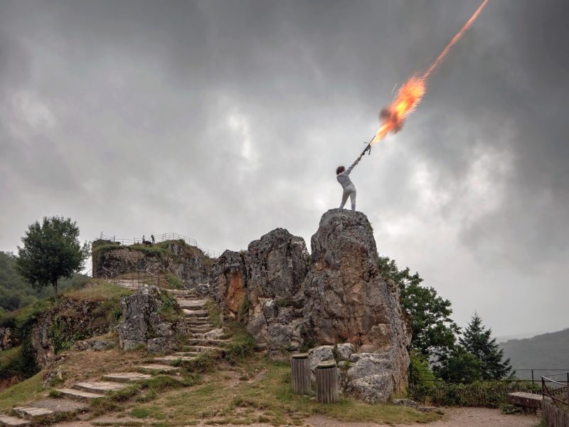 Woman standing on top of rock formation, shooting weapon into the sky. Gloomy day.