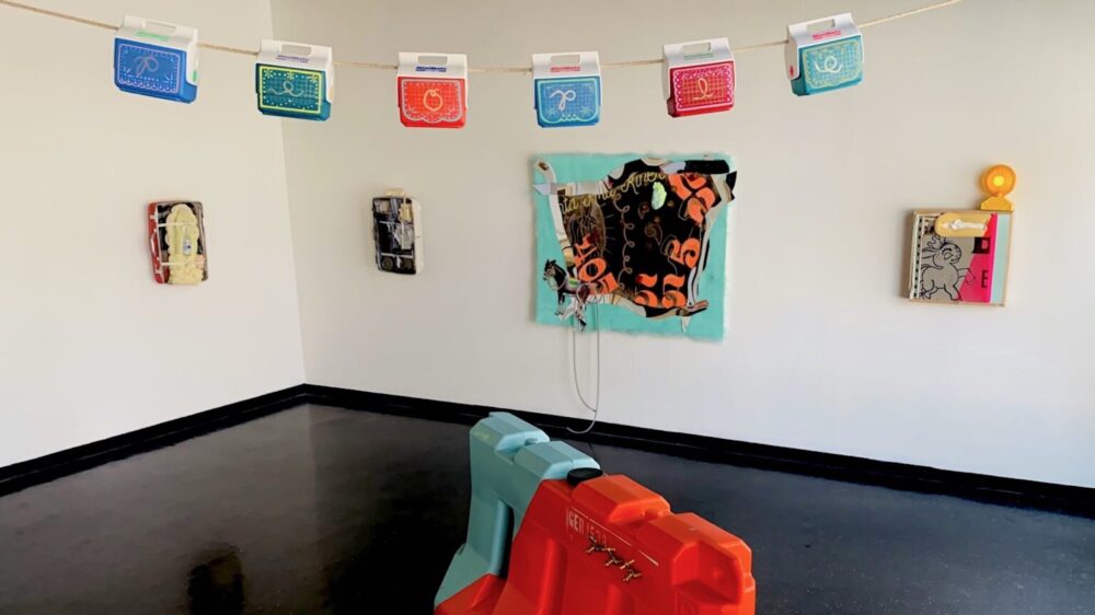 Multi-colored small ice boxes hang vertically from a rope across the gallery with paintings on wall behind and a colorful road barrier underneath.
