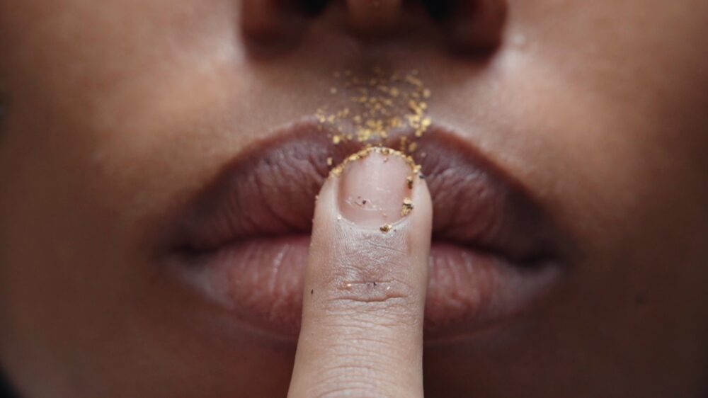 finger with gold dust on it press against the lips of a mouth.