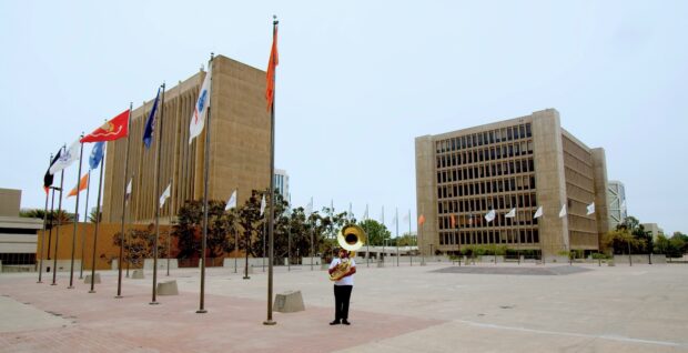 a tube player stands at the edge of a plaza that is lined with flagpoles with two large buildings in the background.