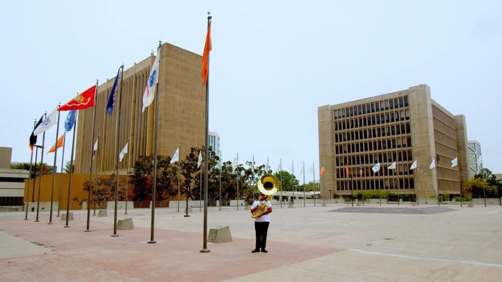 tuba player standing on plaza lined with flag poles with two large buildings in background