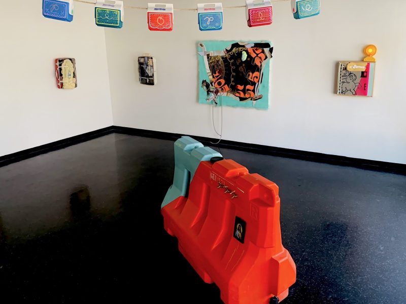 colorful ice coolers hang across a rope toward the top of the image, with four paintings in the backdrop and a large colorful street barrier in the foreground that presentbthe art installation of artist LORENA OCHOA.