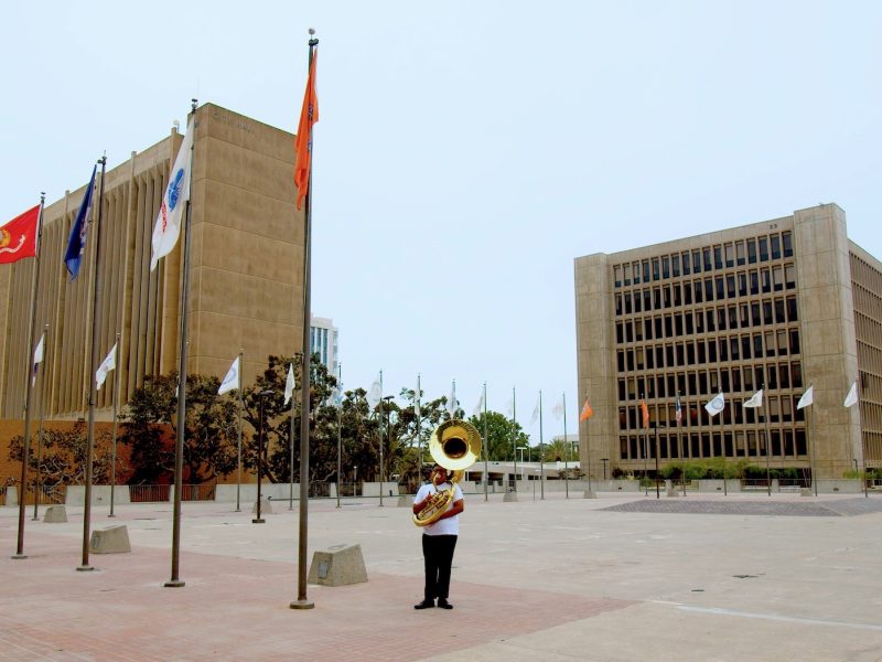 a tube player stands at the edge of a plaza that is lined with flagpoles with two large buildings in the background.