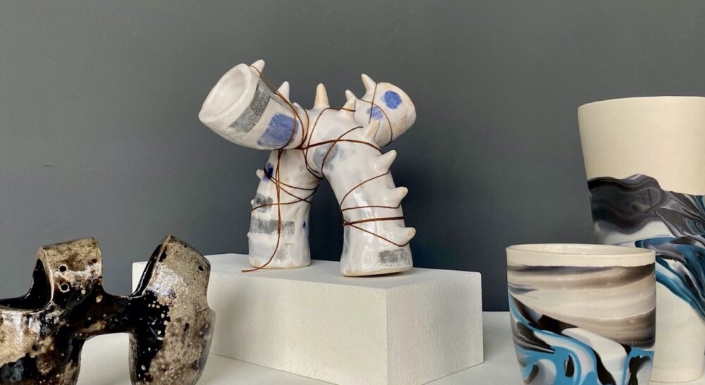 four multi-colored ceramics forms sit on white gallery pedestals in front of a gray painted wall.