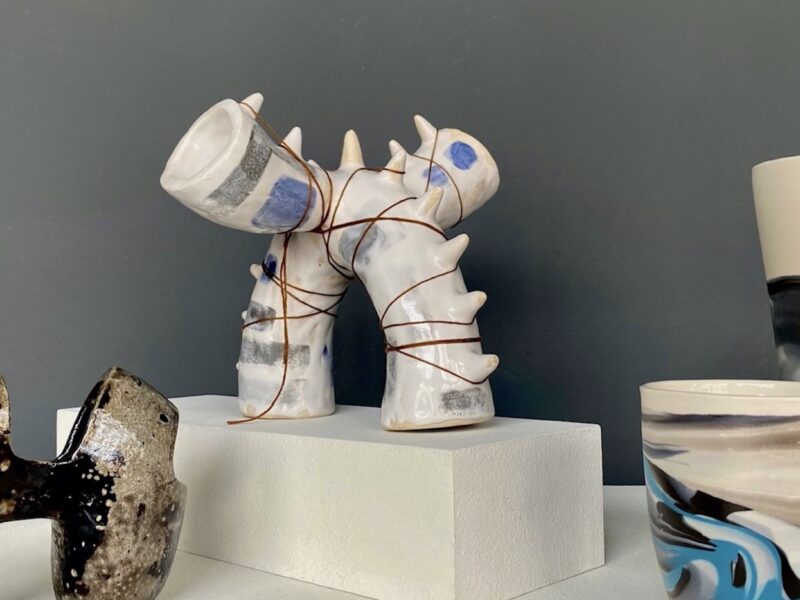 four multi-colored ceramics forms sit on white gallery pedestals in front of a gray painted wall.
