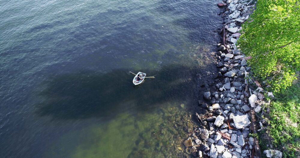 Aerial view from directly above of woman in row boat with a rocky shoreline to the right of image.