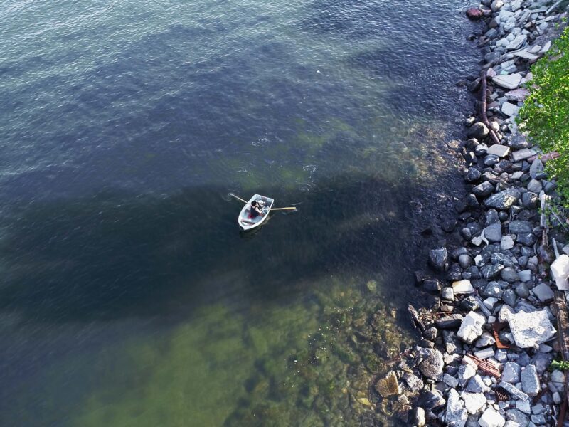 Aerial view from directly above of woman in row boat with a rocky shoreline to the right of image.