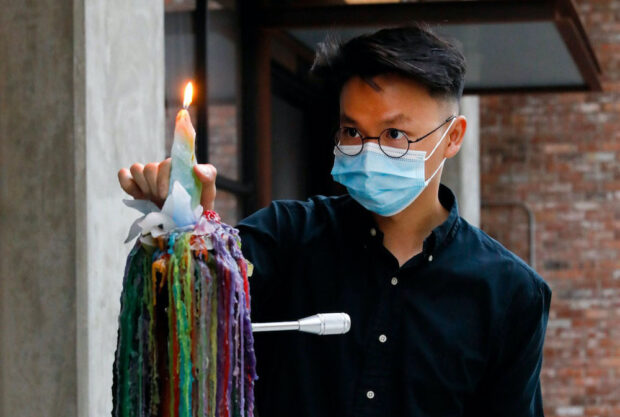 Image of a man standing behind colorful melted candles. He is wearing a surgical mask and has glasses on.