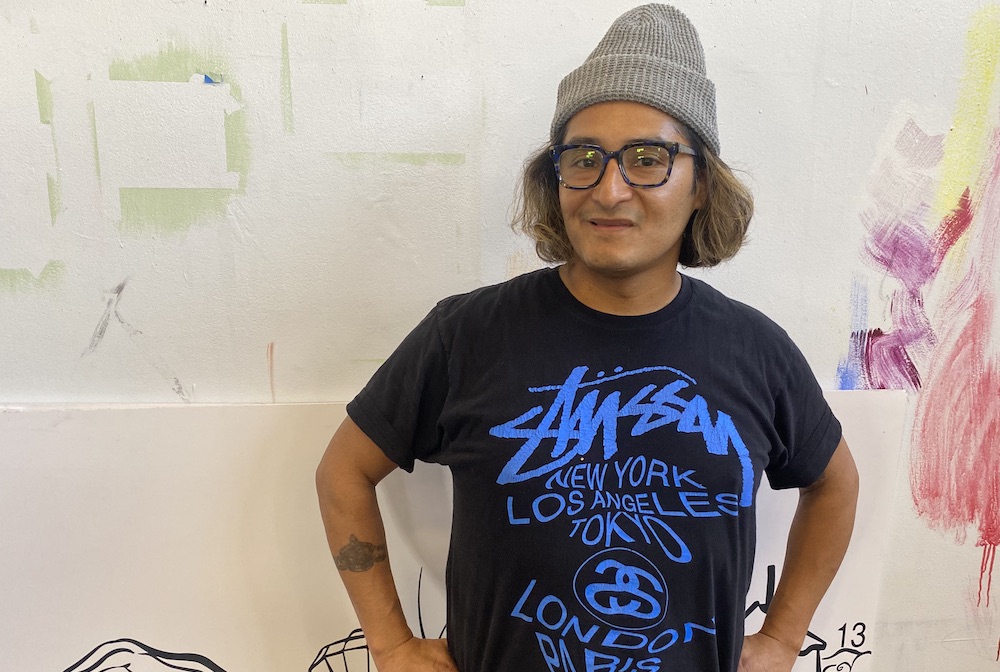 Image of a male with a beanie, brown hair, glasses and black t-shirt. Hands are on his hips, he is standing in front of a white wall with multiple brush stokes on top of it.