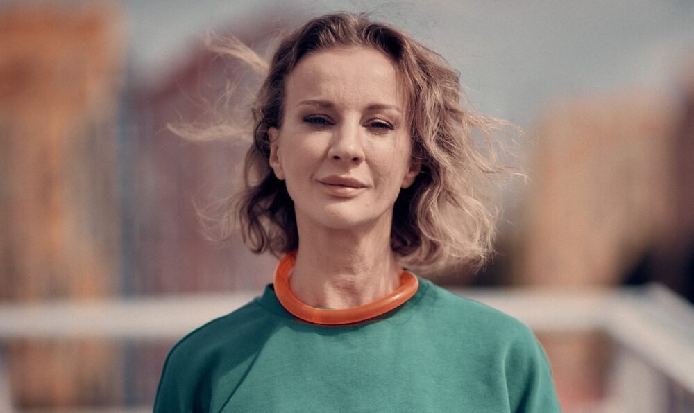 Image of artist Diana Berg, blonde hair wearing an orange short with green sweater on top.
