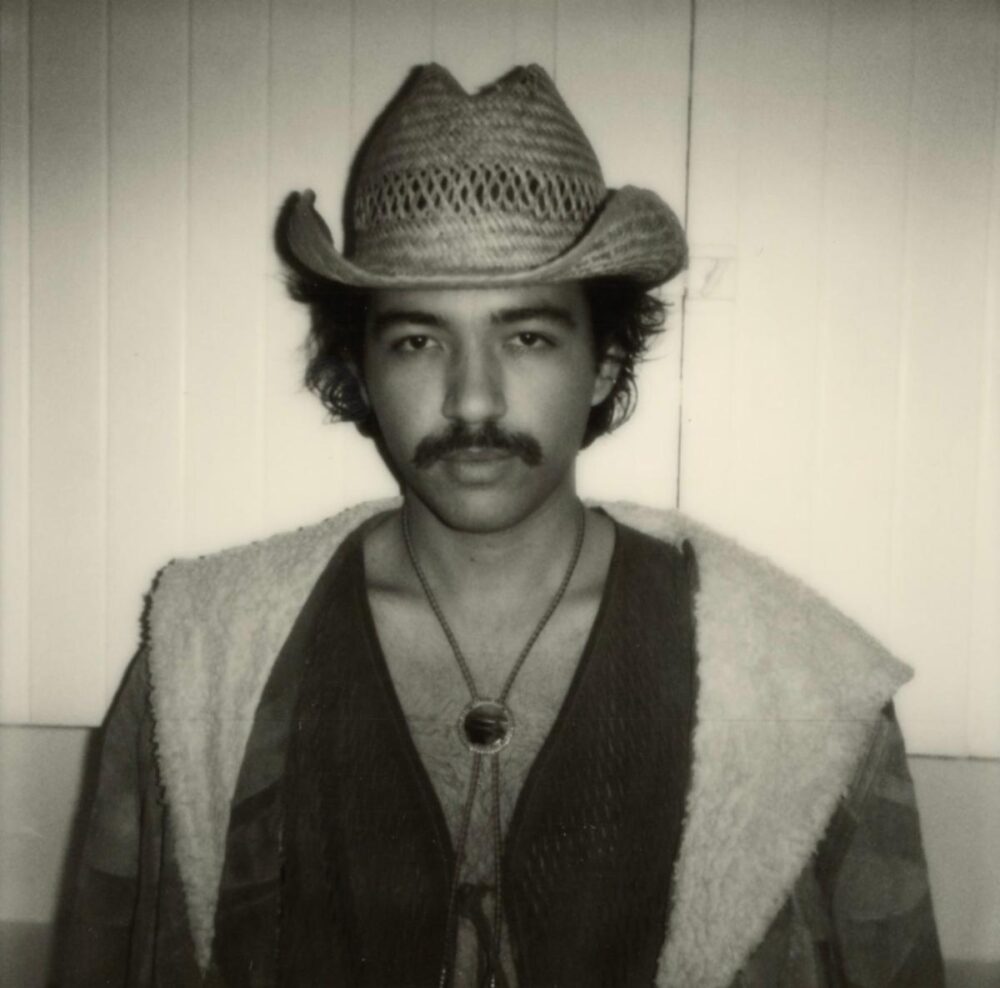 Black and white image of artist Sean Sarmiento, dark hair with mustache, wearing cowboy style hat, black shirt with lighter colored vest. 