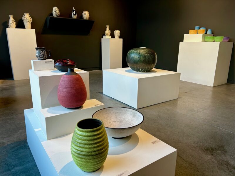 a gallery space with white pedestals holding a variety of ceramic and glass vases and bowls, with a black wall in the background