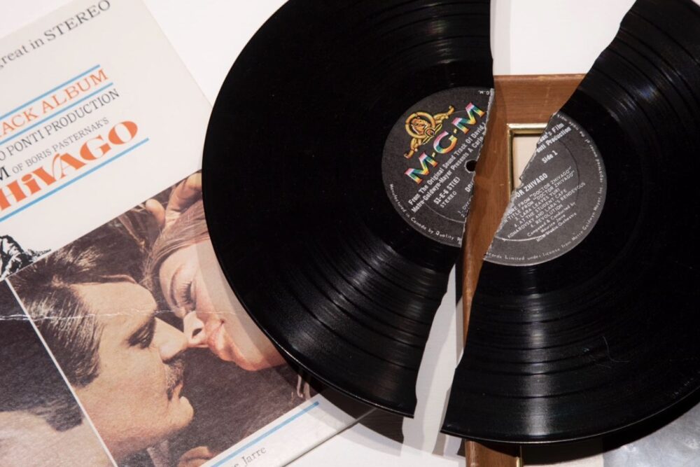 in the background is a partially seen album cover which picture a couple appearing to be ready to kiss. laying on top of the cover is a broken in half black LP record.
