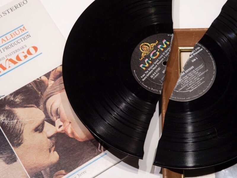 in the background is a partially seen album cover which picture a couple appearing to be ready to kiss. laying on top of the cover is a broken in half black LP record.