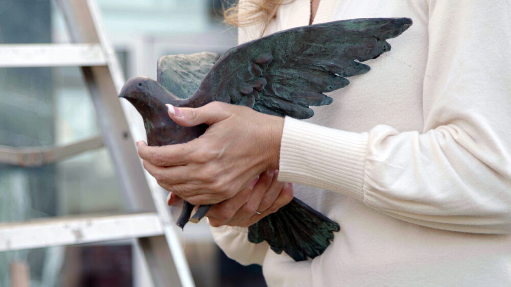 close up image of a woman (Helena Odeh), who is wearing a cream colored sweater, and holding a bronze dove in her hands