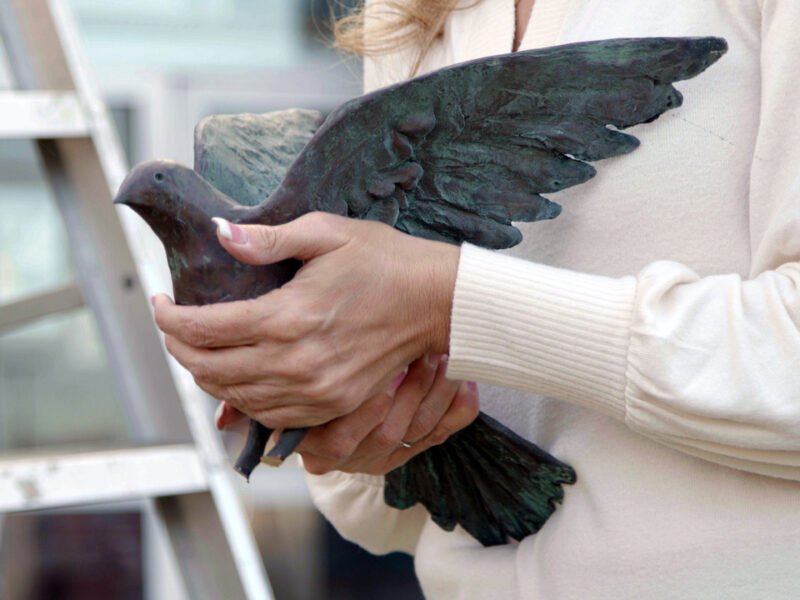 close up image of a woman (Helena Odeh), who is wearing a cream colored sweater, and holding a bronze dove in her hands