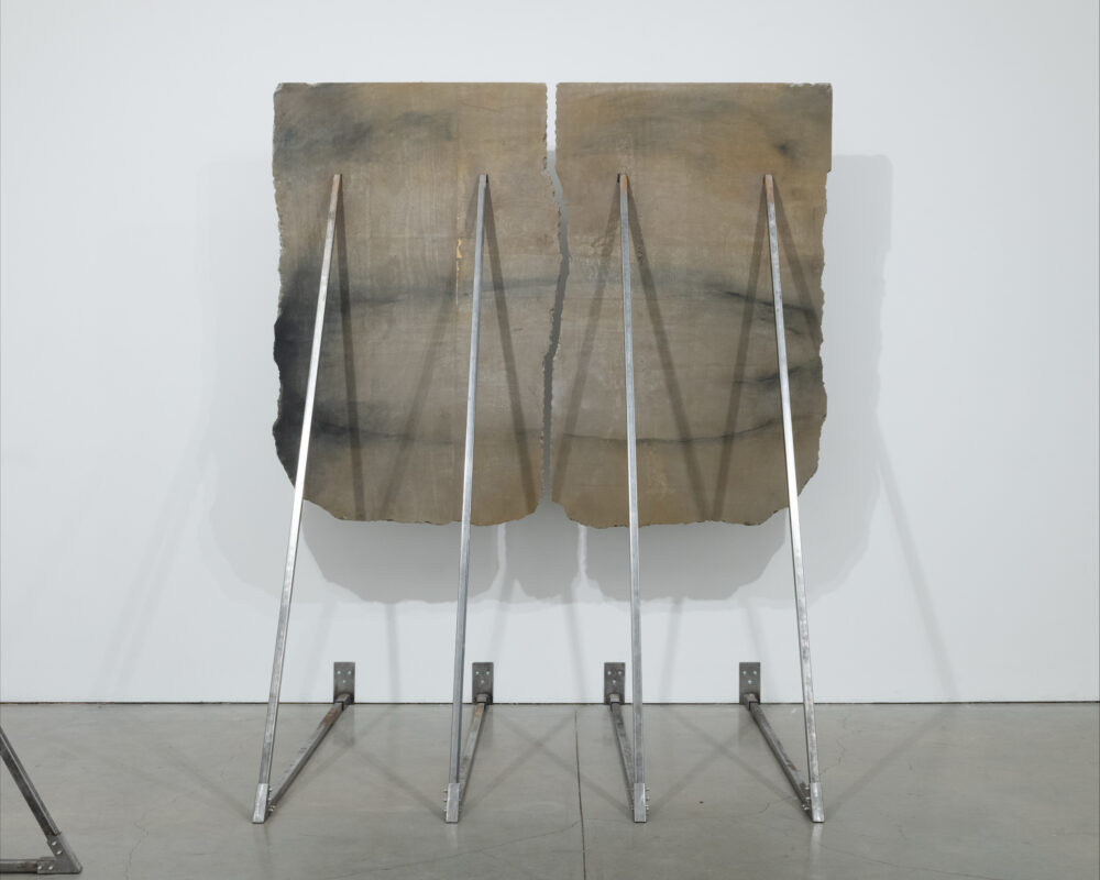 Two large vertical concert panels being pushes and supported halfway up a white gallery wall by metal triangle support posts.