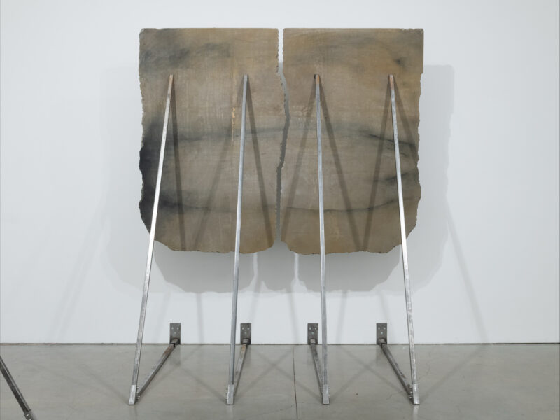 Two large vertical concert panels being pushes and supported halfway up a white gallery wall by metal triangle support posts.