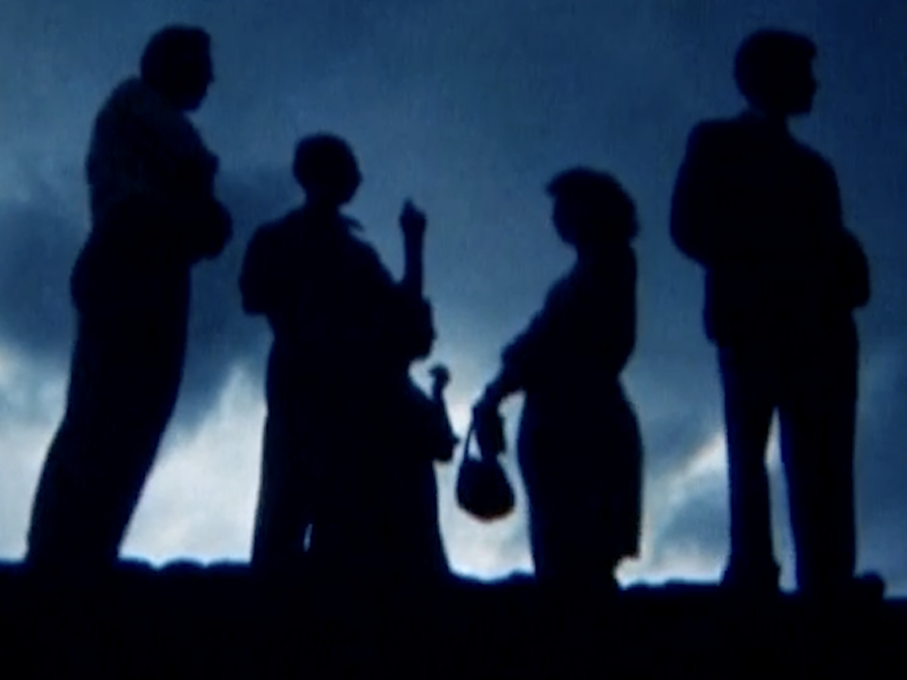 A blue-toned image shows the dark silhouettes of four people standing against a softly illuminated, cloudy background. One figure gestures with a raised hand, while another carries a small handbag. The hazy lighting and deep shadows create a cinematic, atmospheric scene.