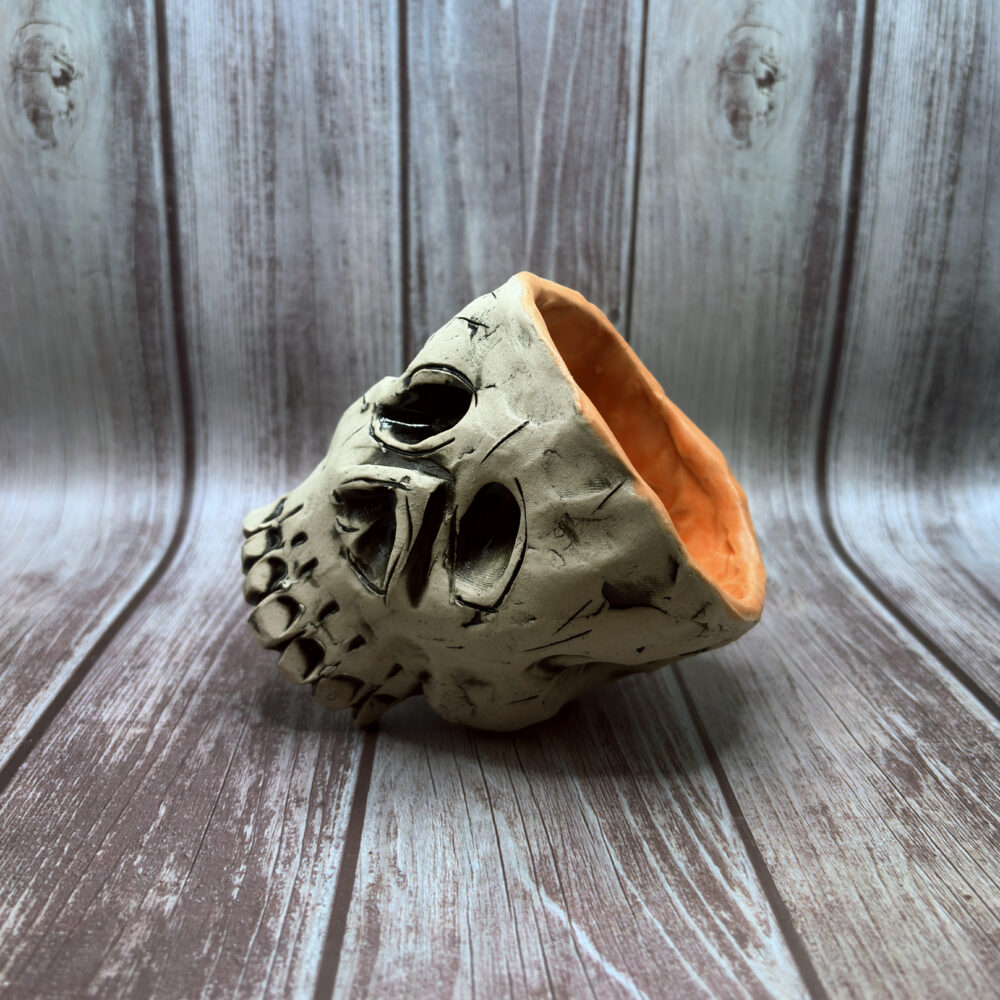 A hand-built ceramic sculpture resembling a stylized skull, with a rough gray surface, carved dark facial features, and an open back revealing a warm orange interior, photographed against a soft blue-gray background.