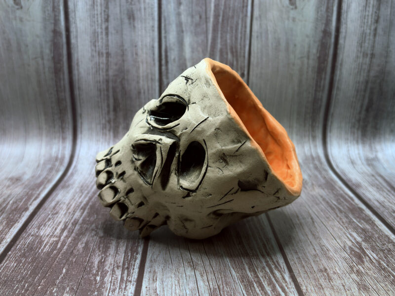 A hand-built ceramic sculpture resembling a stylized skull, with a rough gray surface, carved dark facial features, and an open back revealing a warm orange interior, photographed against a soft blue-gray background.